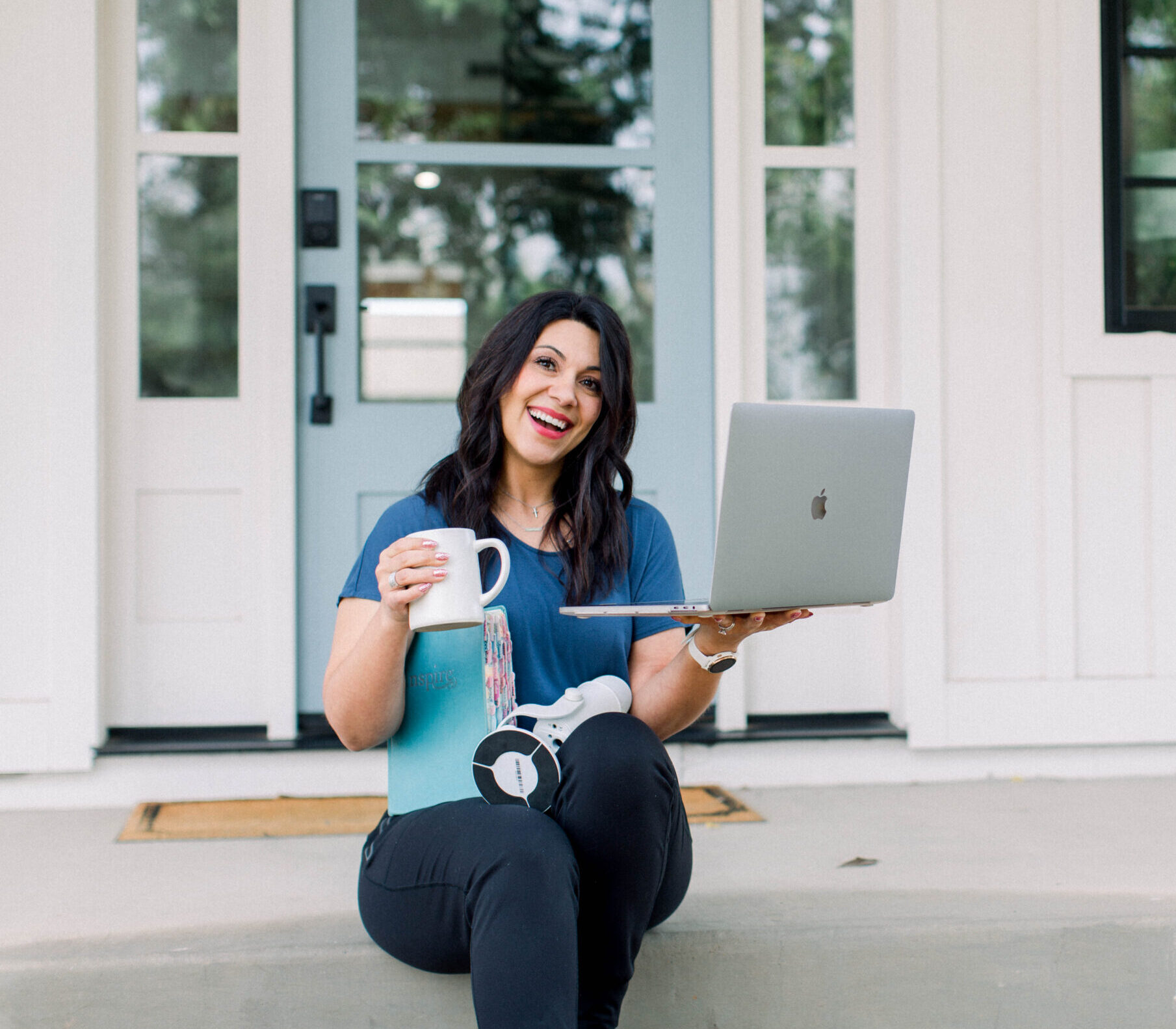 a smiling brown haired woman sitting on her front porch steps holding her coffee cup and laptop in her hands. Her day planner and podcast microphone sit in her lap.