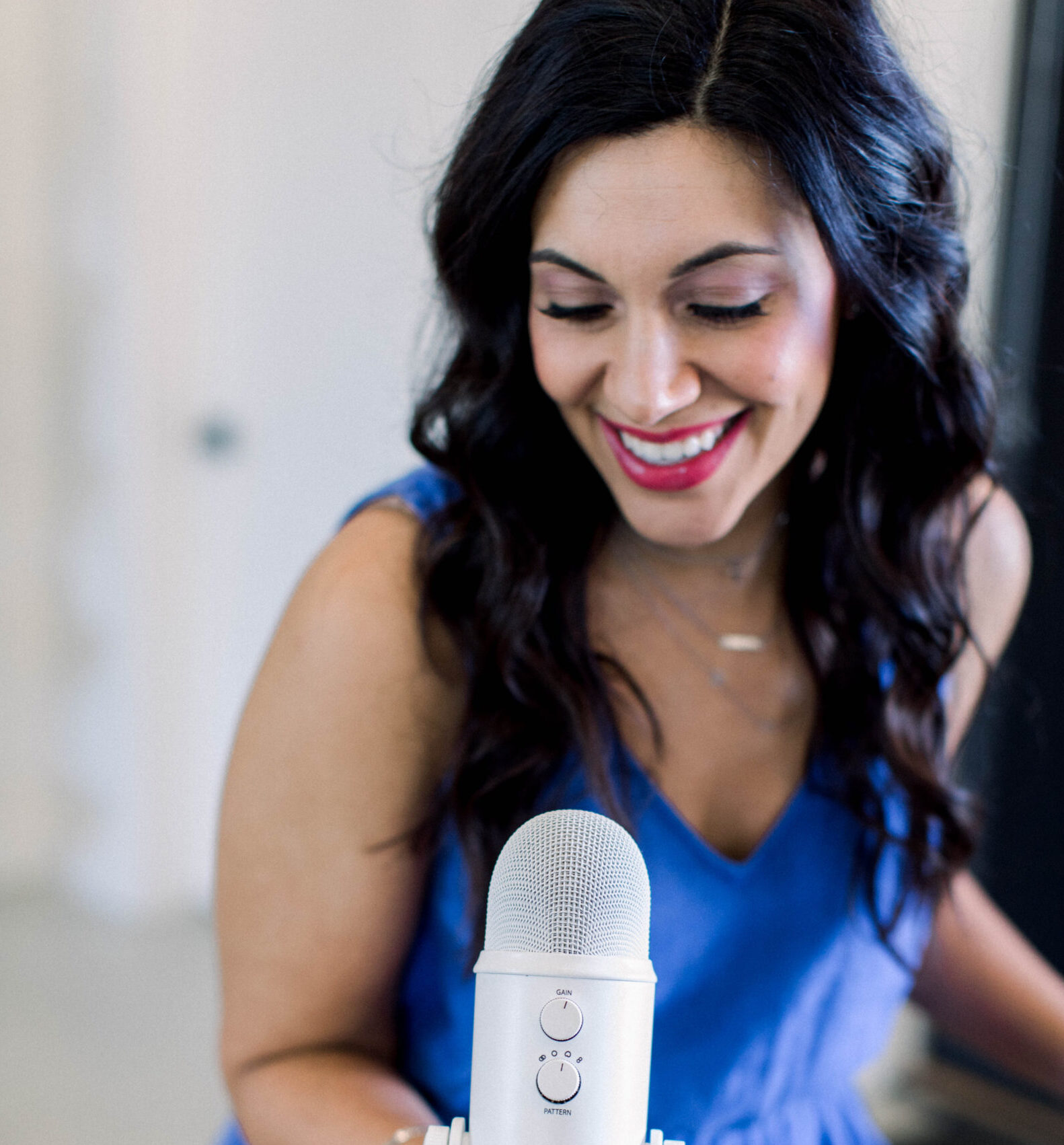 Image is a woman with dark hair smiling while looking at a podcast microphone