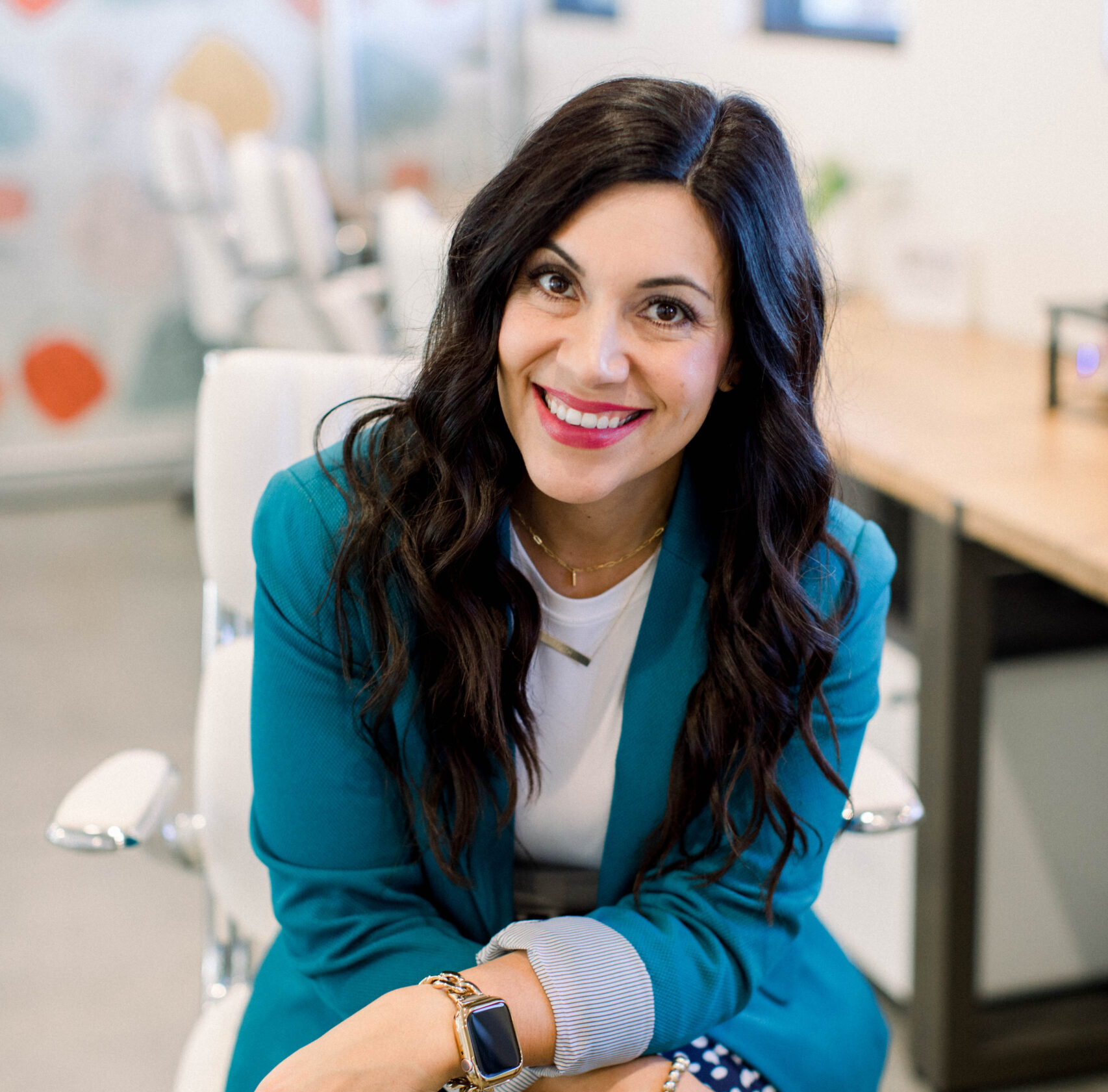 Smiling brown haired woman sitting on a white chair with arms crossed in her lap, she is leaning forward.