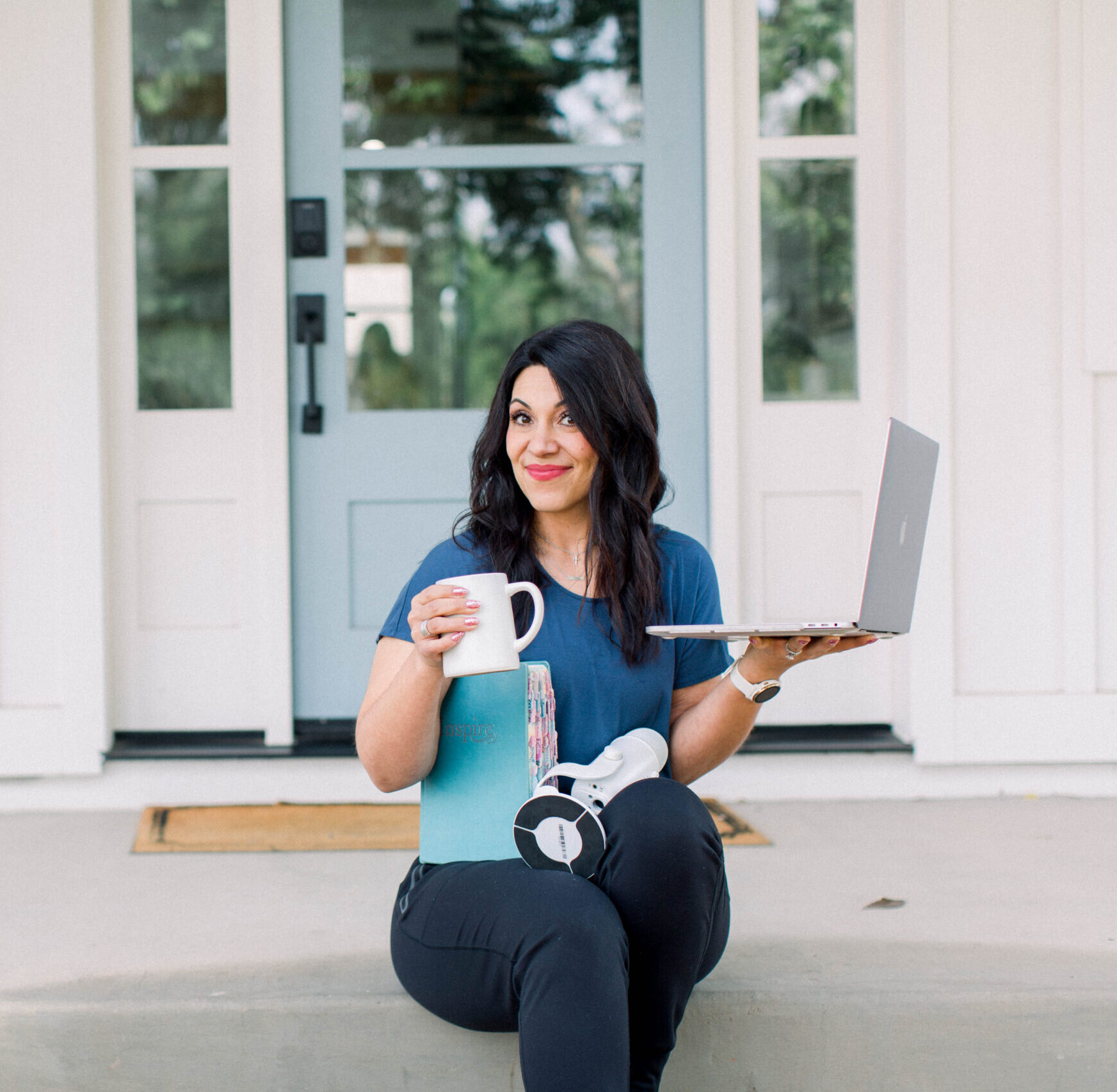 Woman is seated on steps with a coffee cup, notebook, and laptop in her hands.