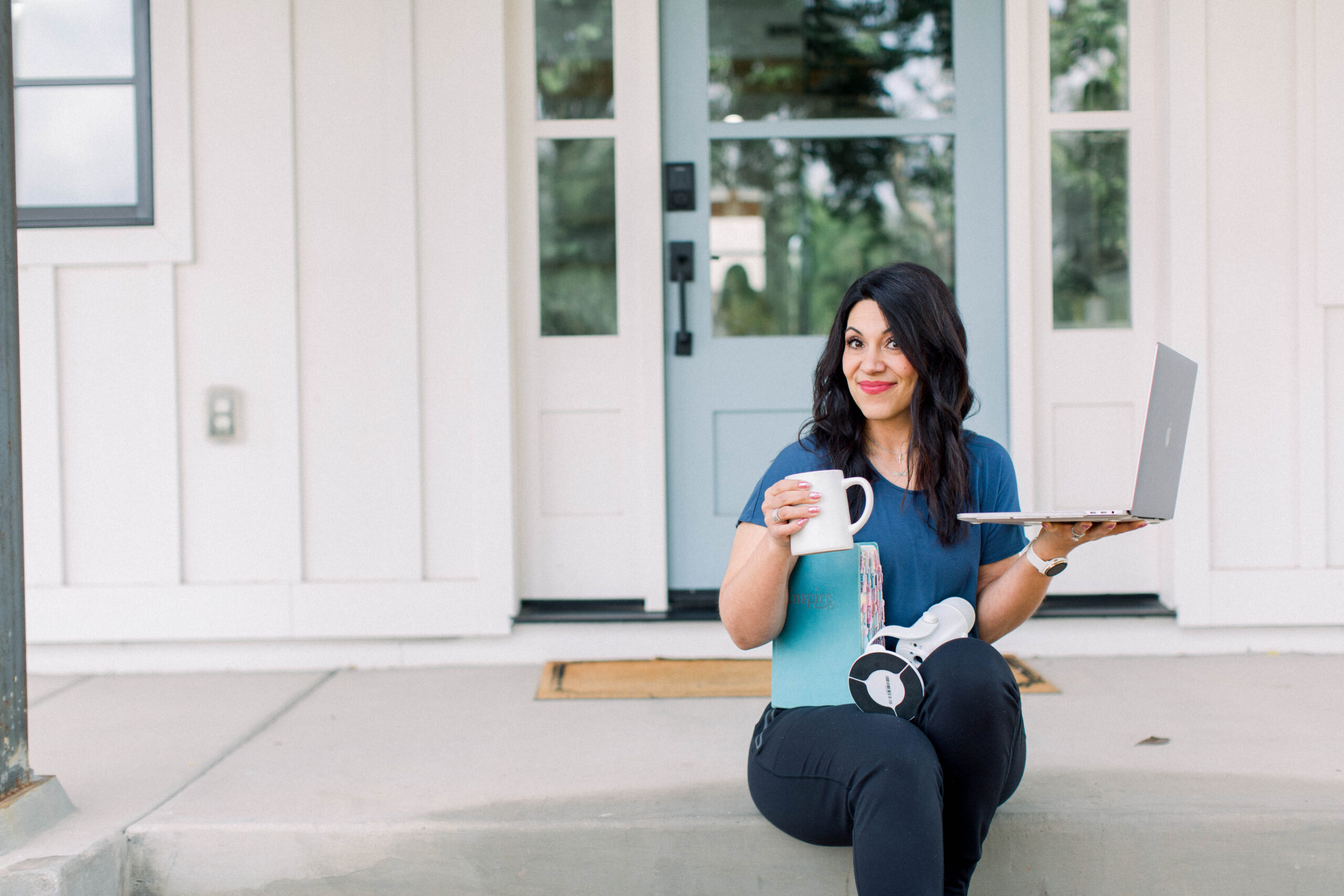 Woman is seated on steps with a coffee cup, notebook, and laptop in her hands.