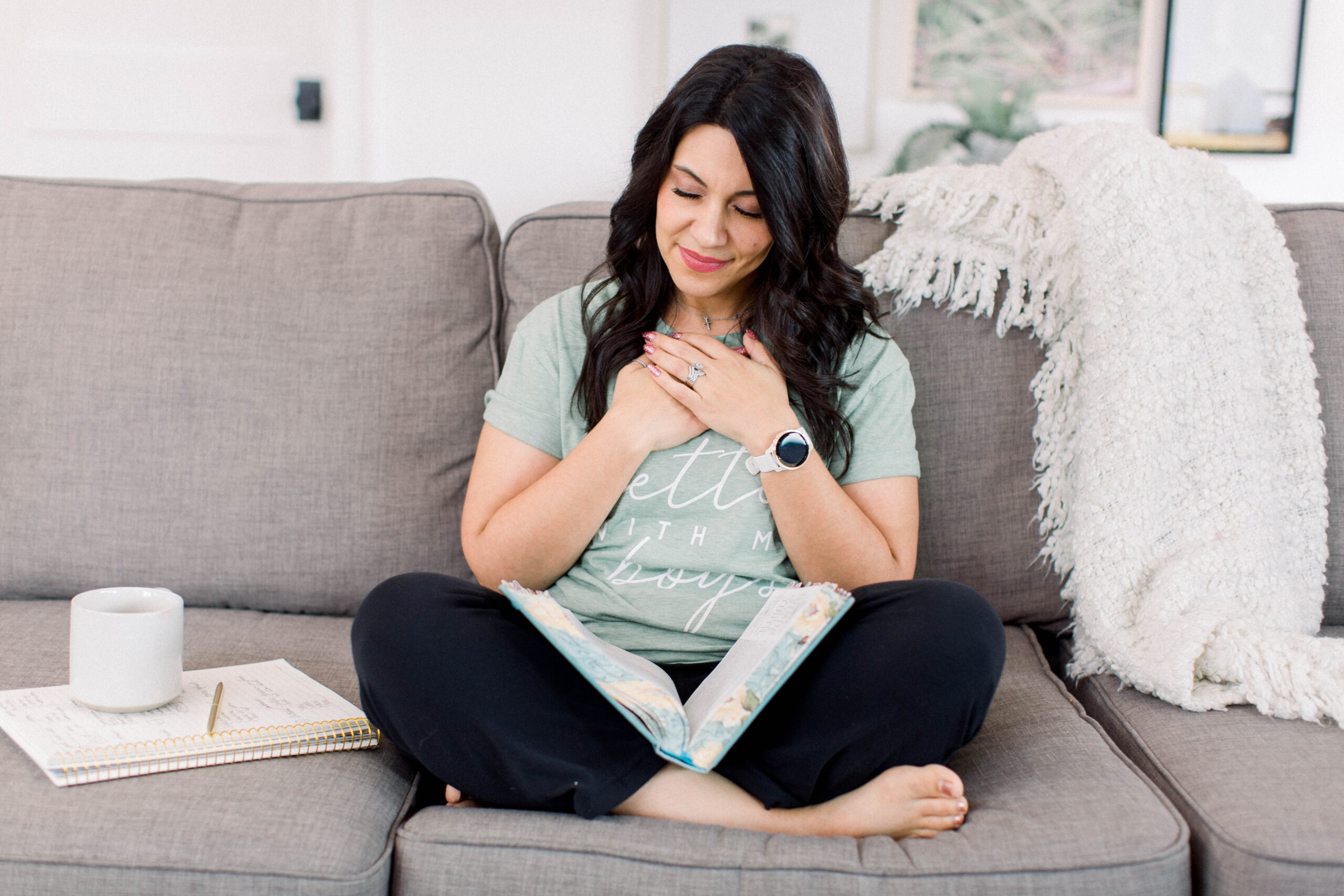 brown haired woman sitting on her couch reading her Bible which is in her lap. Her coffee mug is beside her.  Her hands are clasped together on her chest. 