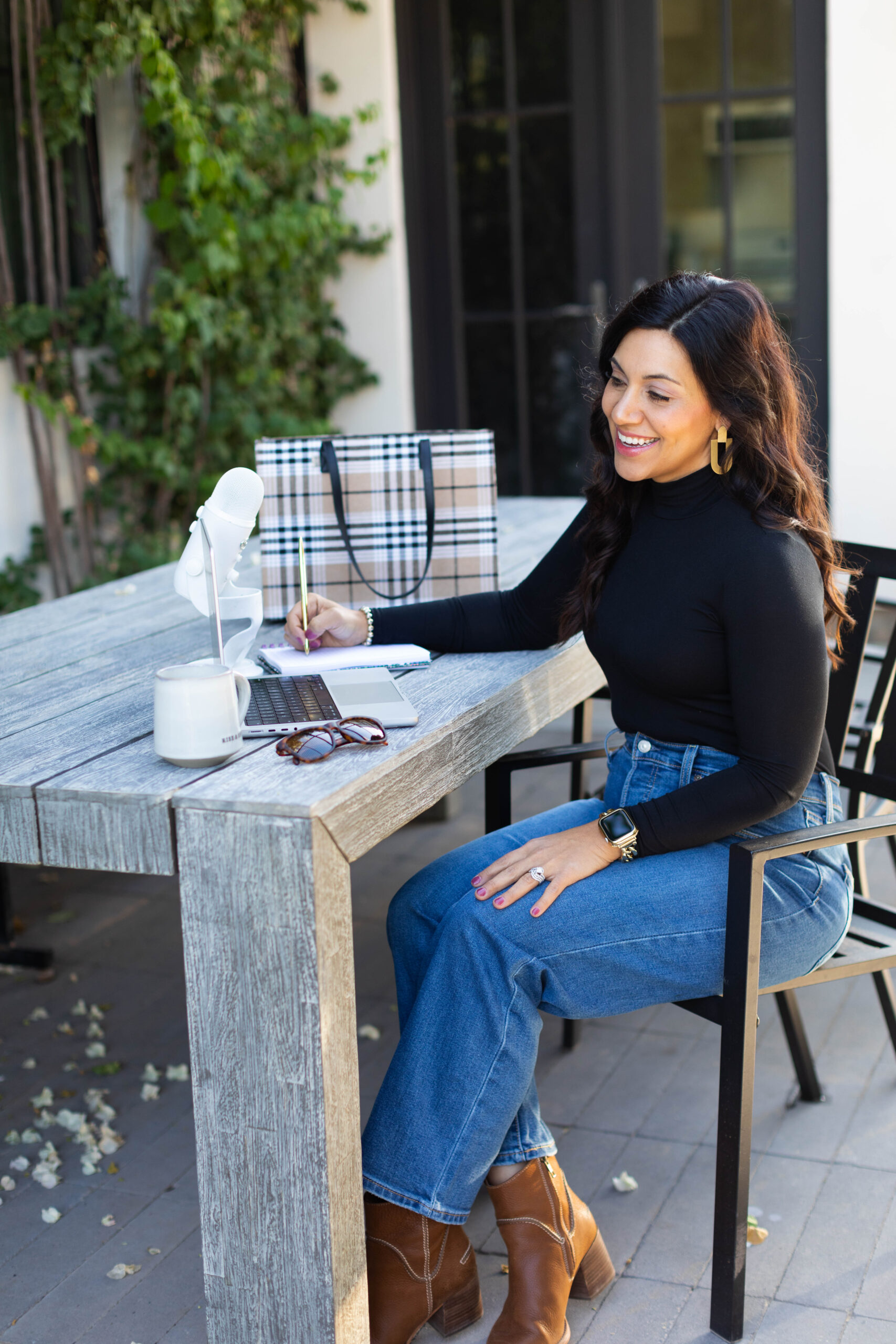 Image is a woman in a black shirt with jeans smiling while sitting at a table with a laptop and microphone. 