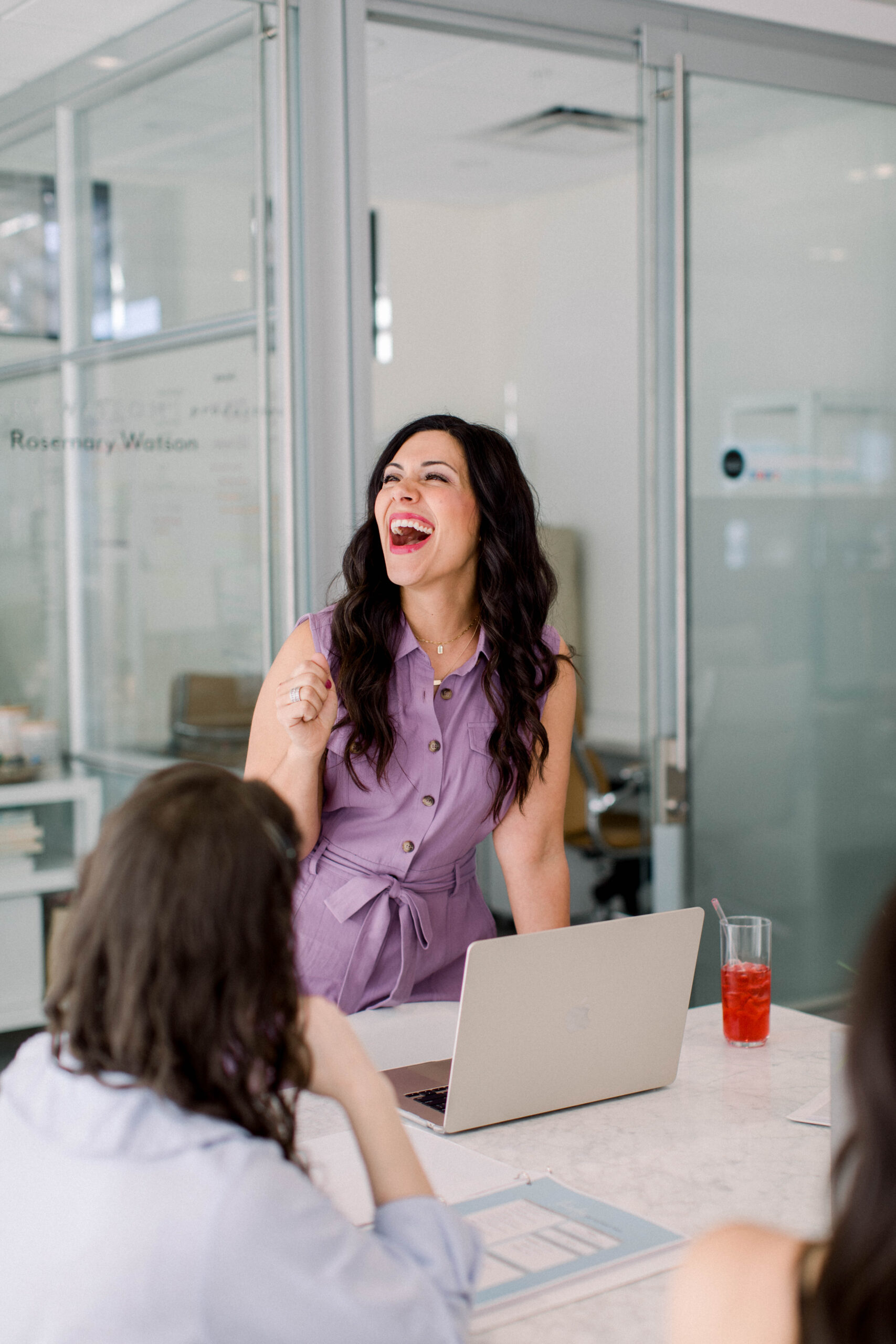 Image is a dark haired woman smiling while standing next to her computer. 