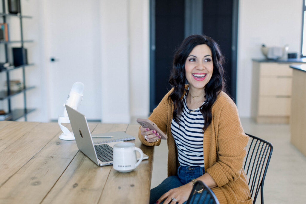 brown haired woman smiling as she holds her cell phone in her hand. She is sitting at her desk with her laptop and white podcast microphone, Stefanie Gass