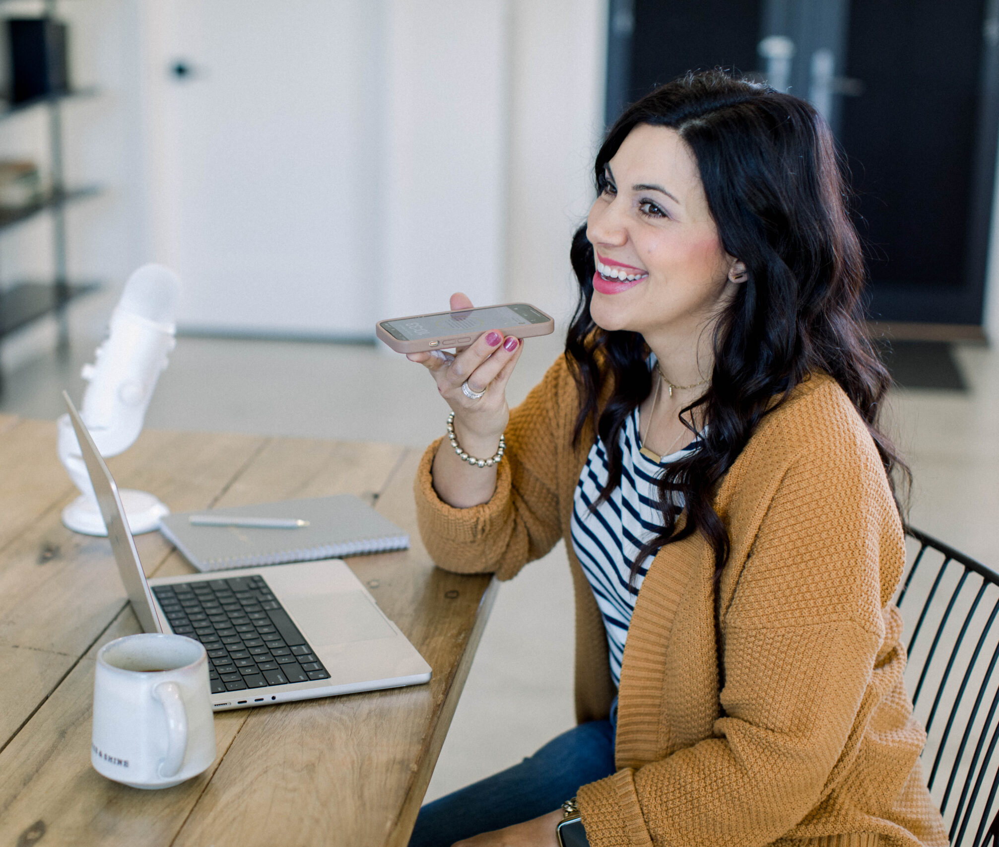 brown haired woman smiling as she talks into her cell phone. She is sitting at her desk with her laptop and white podcast microphone,