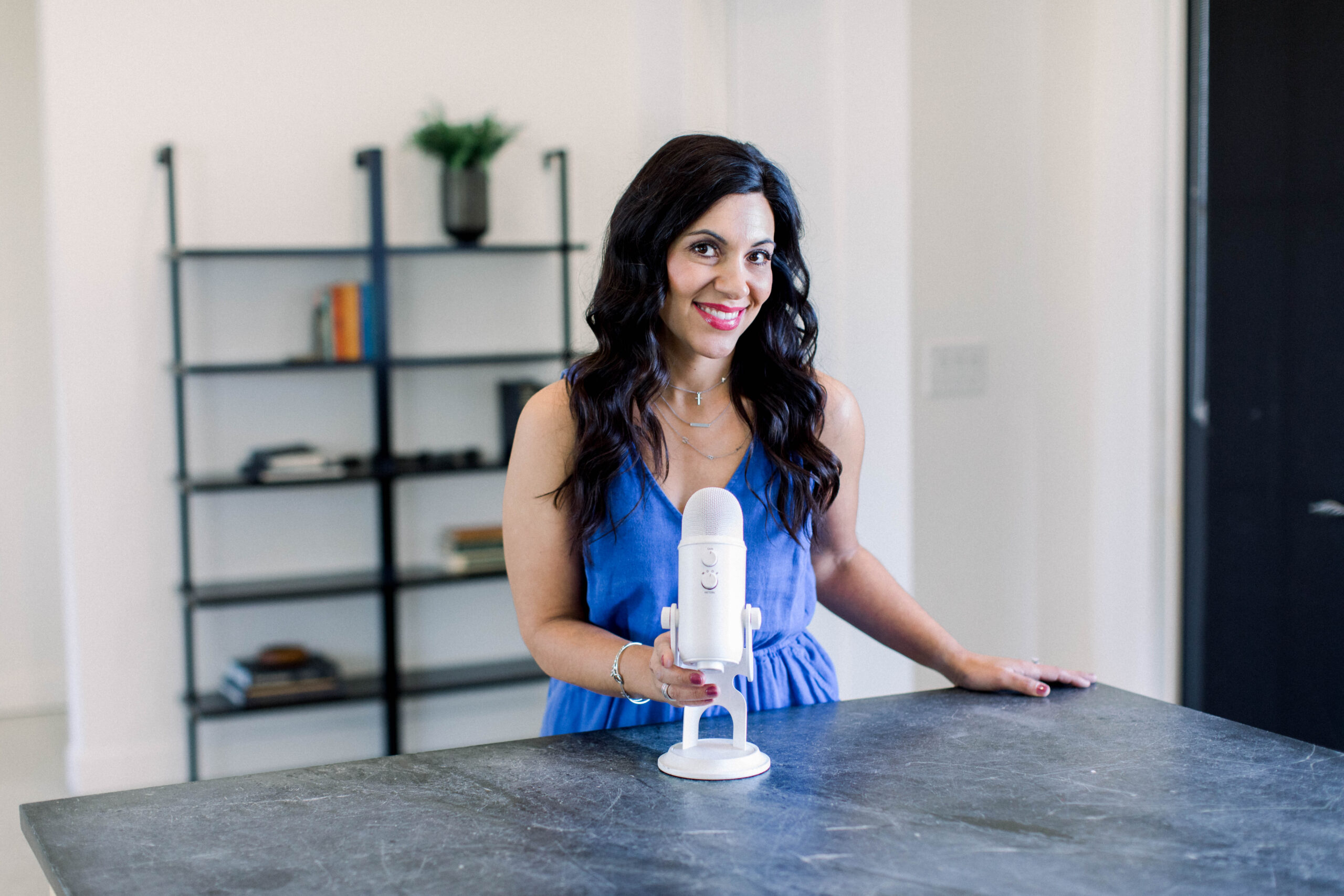 brown haired woman smiling as she stands at a black countertop with her white podcast microphone in her hand.