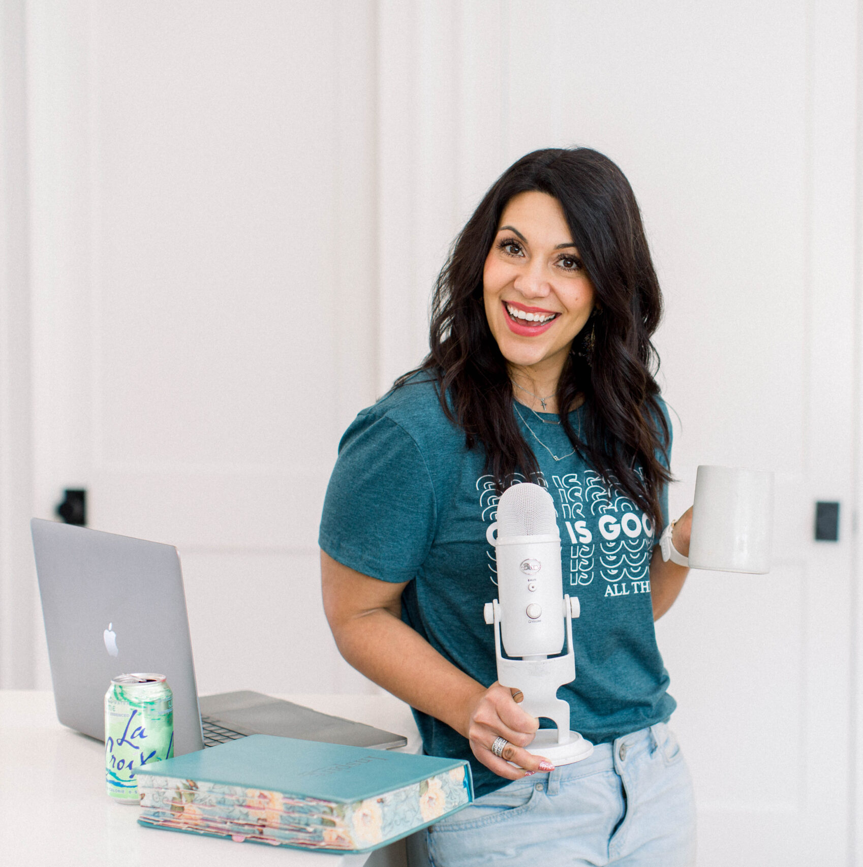 brown haired woman smiling as she holds her white podcast microphone in her right hand. Her laptop and day planner is on a table beside her