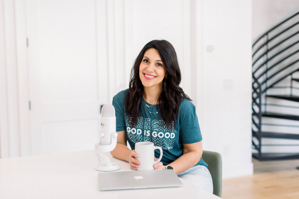 brown haired woman smiling as she sits at a table with her white coffee cup in her hand, her laptop, and white podcast microphone