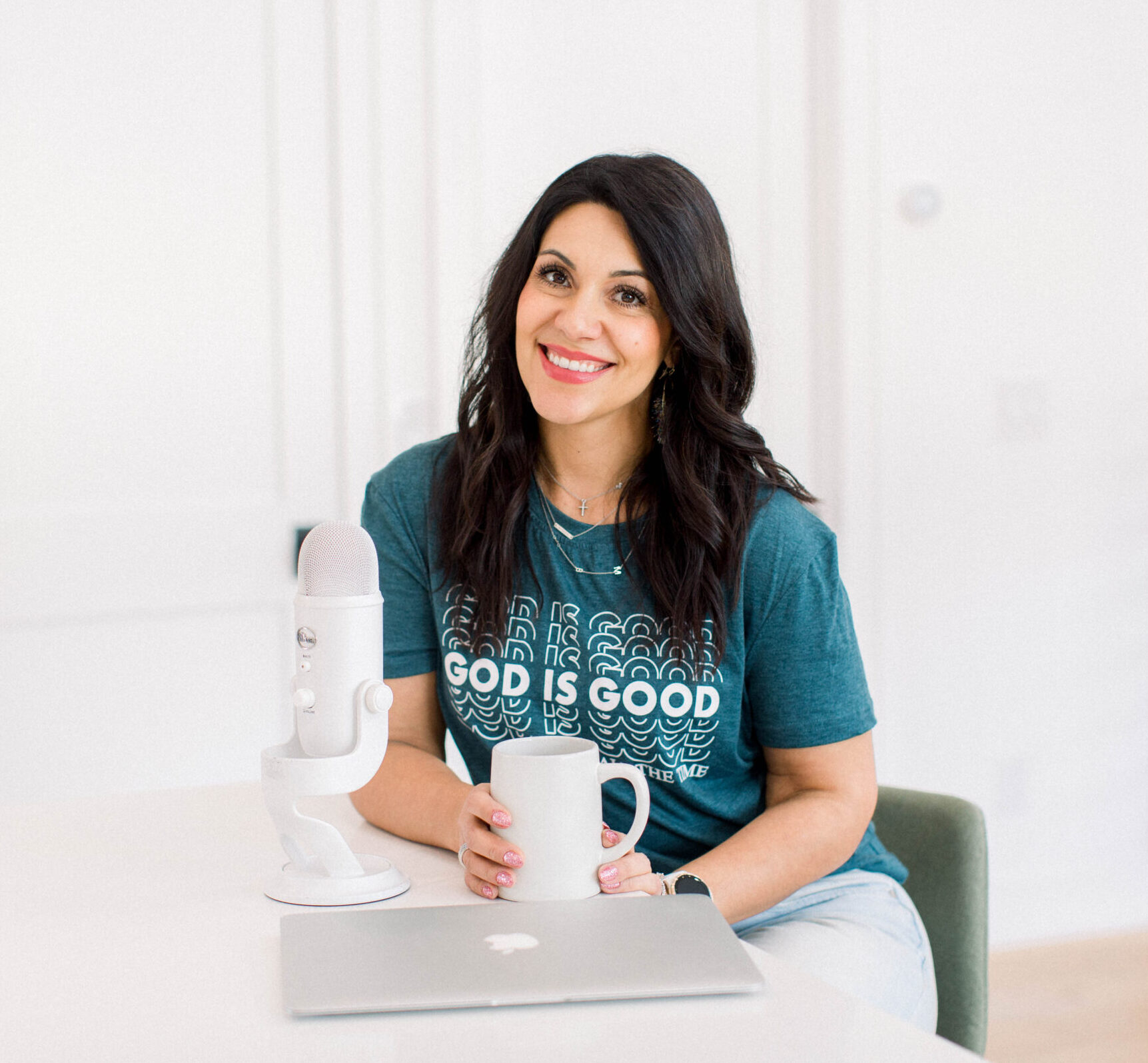 brown haired woman smiling as she sits at a table with her white coffee cup in her hand, her laptop, and white podcast microphone