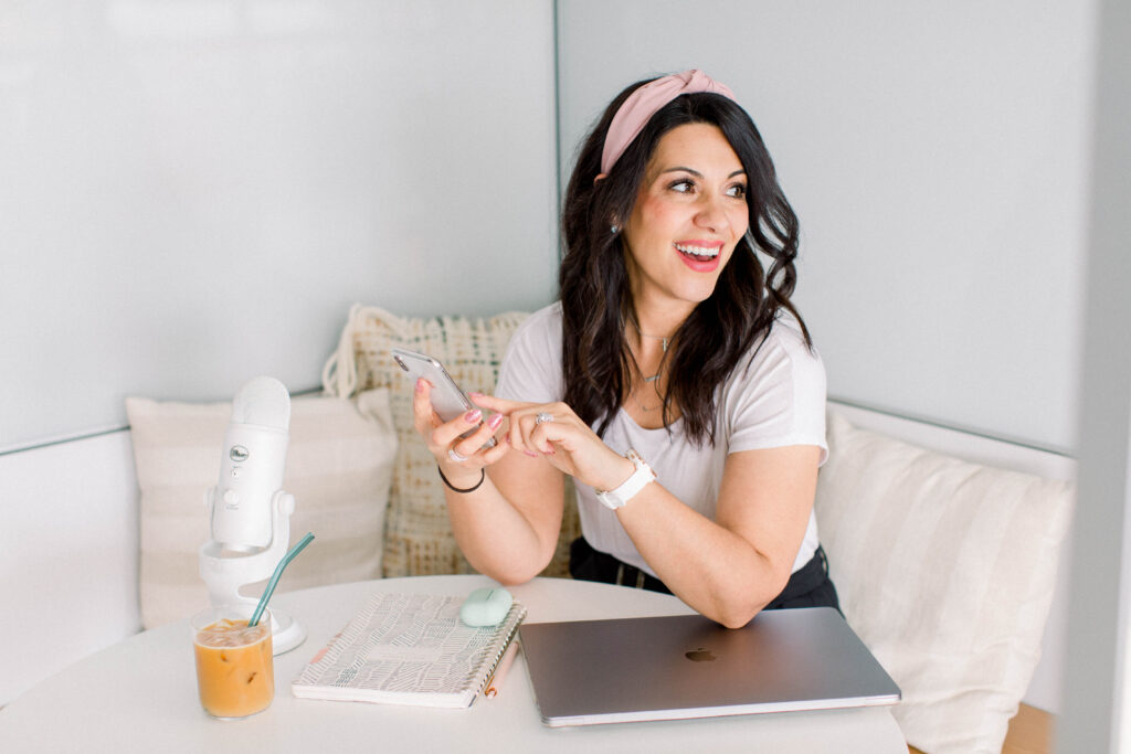 brown haired woman smiling with her cell phone in her hand. She has her laptop and white podcast microphone on the table she is sitting at. Stefanie Gass