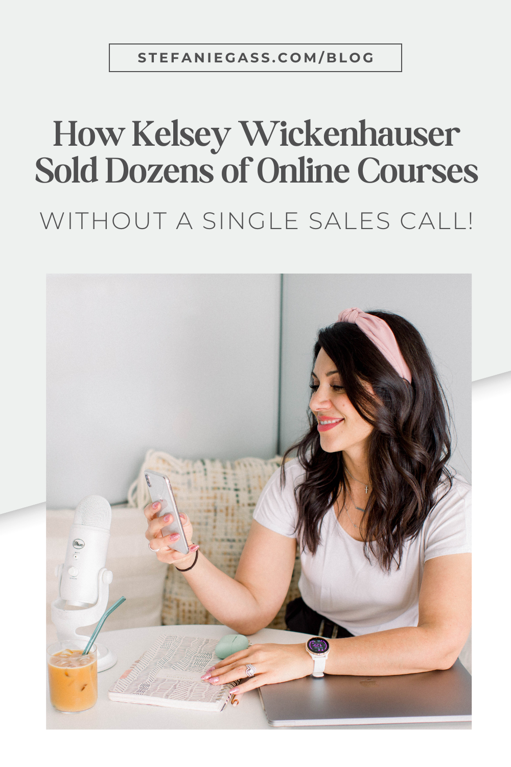 brown haired woman smiling as she looks at her cell phone. She has her laptop and white podcast microphone on the table she is sitting at. Image text reads: How Kelsey Wichenhauser Sold Dozens of Online Courses without a Single Sales Call. Stefanie Gass Blog