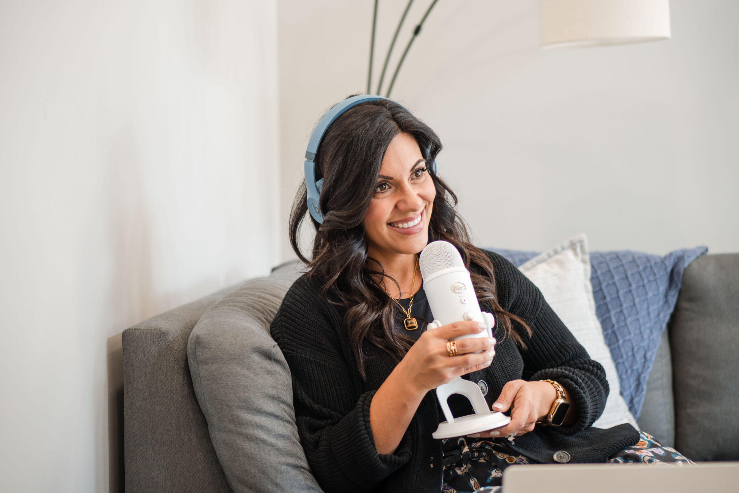 relaxing-on-the-couch-holding-microphone-wearing-headphones-podcasting-smiling-Stefanie-Gass-horizontal The image title is: How-to-Navigate-Podbean-Podcasting-Software-Stefanie-Gass