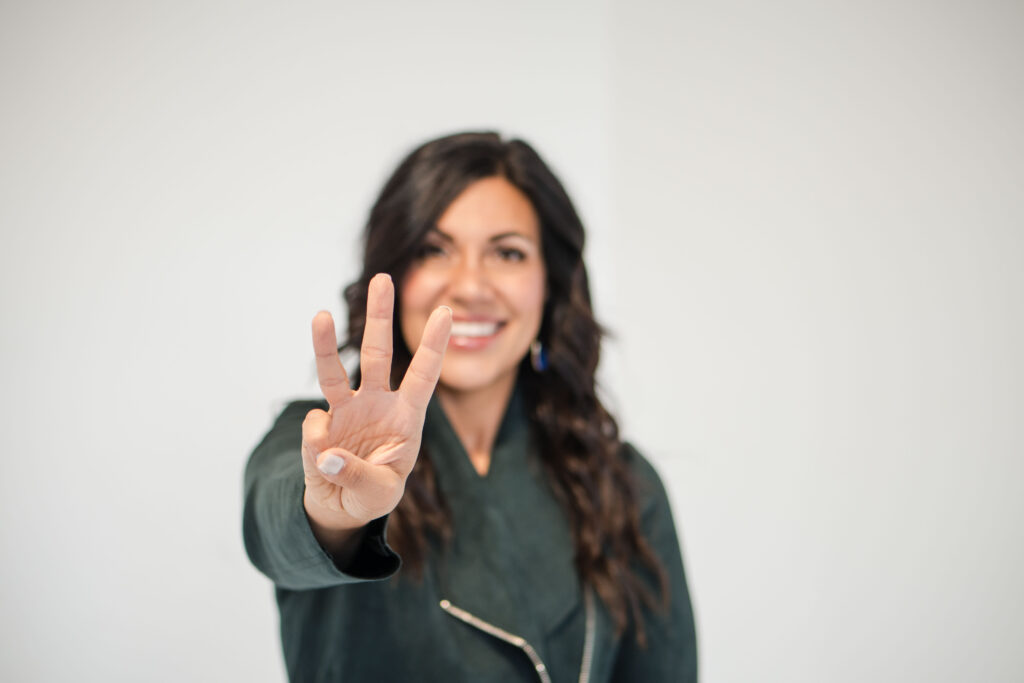 A dark-haired woman is smiling in the background and her face is blurred. In the front of the image, she's holding three fingers to the front. The title of the image is: Build-a-Successful-2025-Business-Plan-with-the-Three C-Stefanie-Gass