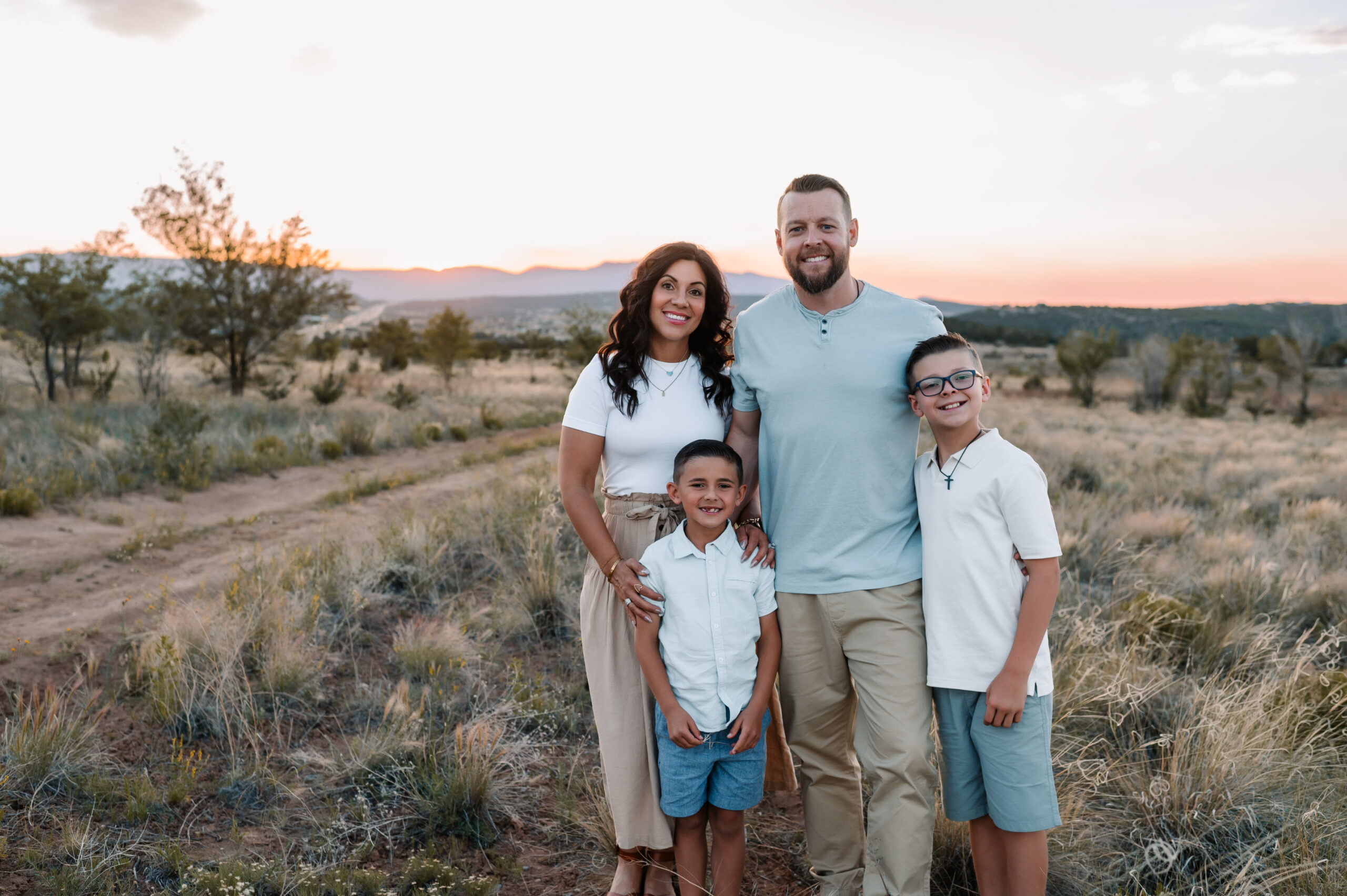 Family of four smiling in a forest. They're standing next to each other, with the adults in the back, and the kids in the front. The two boys and the mom are wearing a white t-shirt, and the dad is wearing a blue t-shirt.