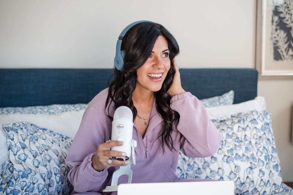 Dark-haired woman sitting in bed wearing a light purple sweater. She's wearing blue headphones and seems focused as she's also holding a white microphone in her hand. The image title is: 10-Reasons-Podcasting-is-the-Best-Way-to-Make-Money-Online-Stefanie-Gass