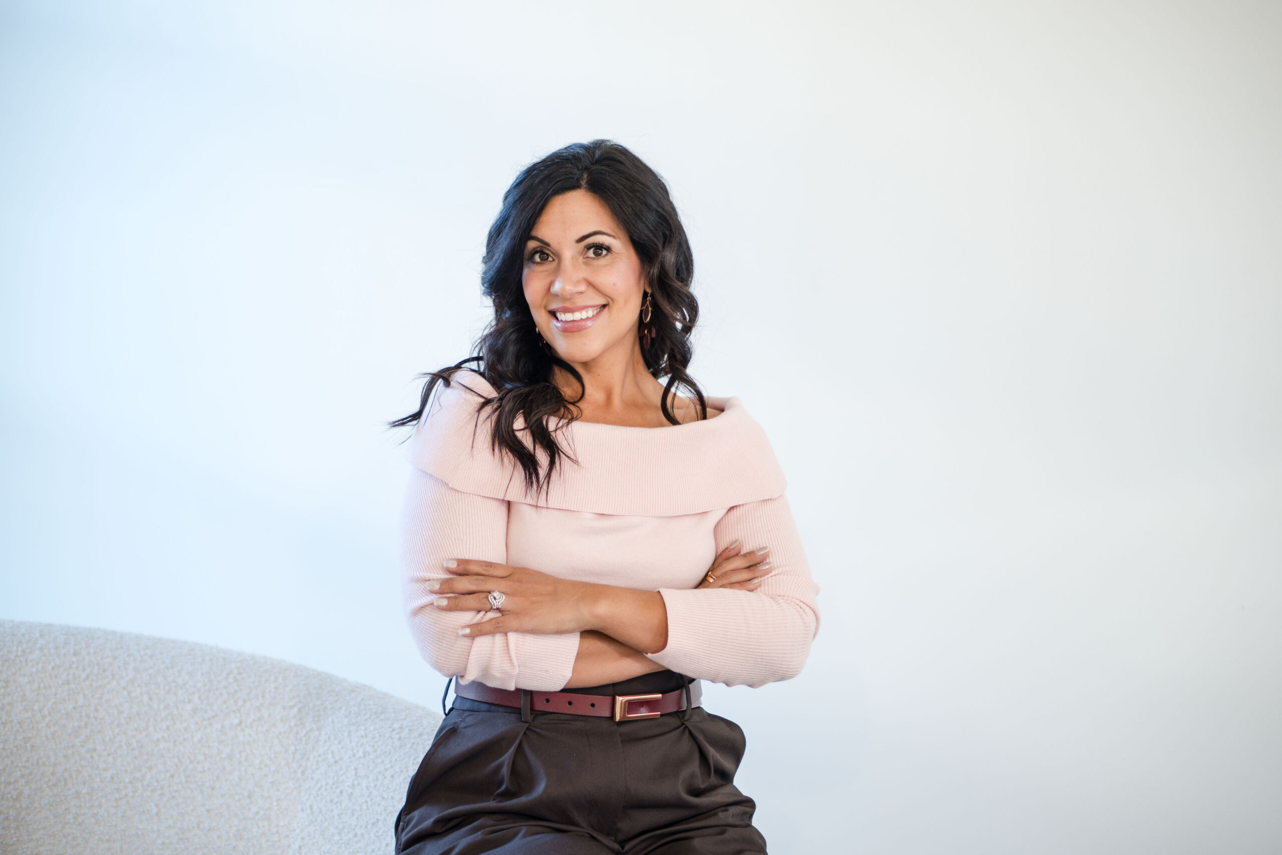 Dark-haired woman standing leaning against a white couch. She's wearing a light pink sweater and brown pants. The image title is: Build-Systems-and-Get-Organized-with-Chelsi-Jo-Moore-New-Year-New-You-Stefanie-Gass