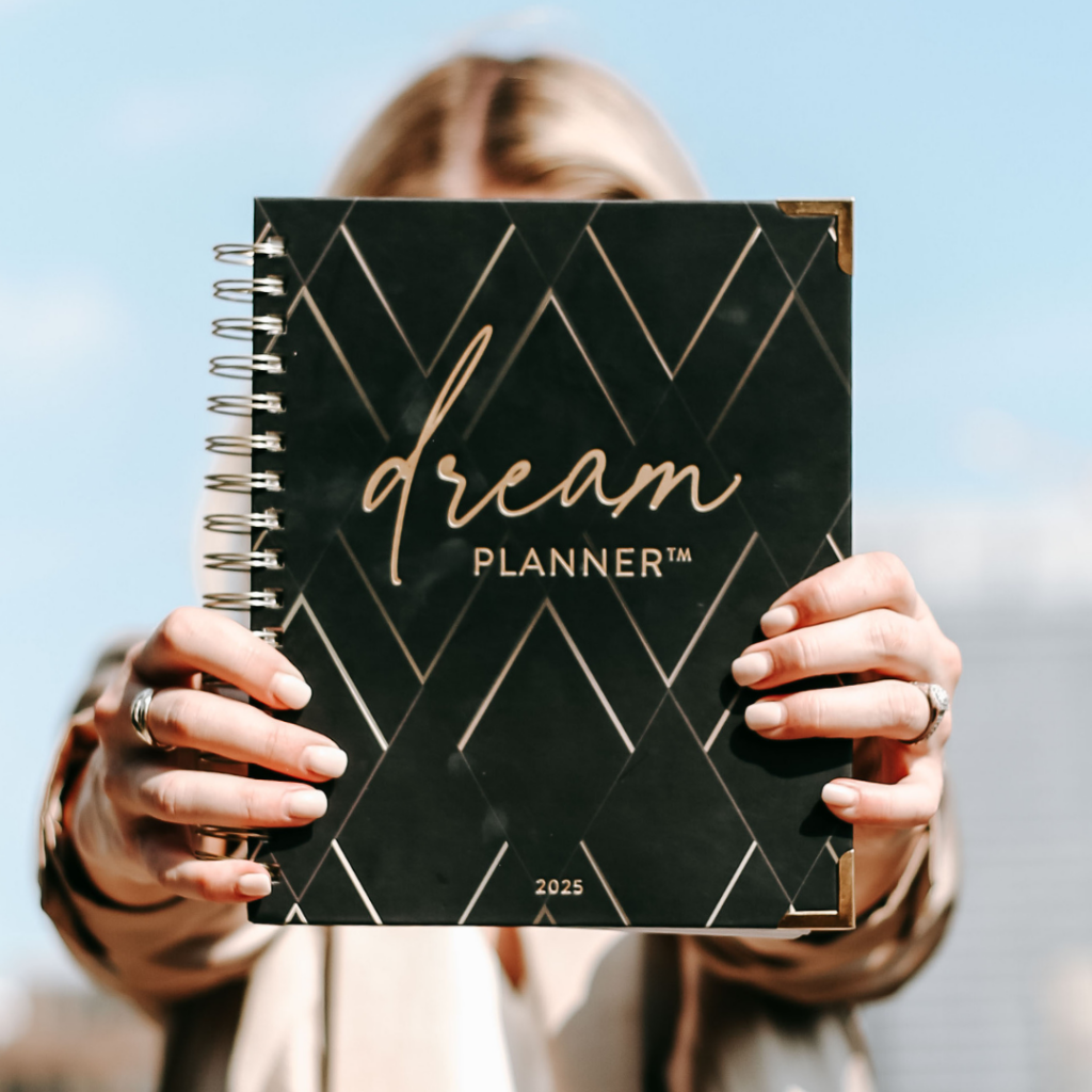 Blonde woman holding up a Horacio Dream Planner in front of her face with a blue sky background.