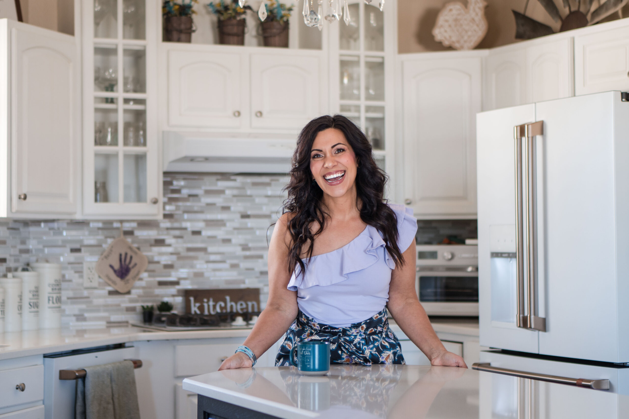 A brunette woman is happy standing in her kitchen. She is wearing a one-shoulder lilac top and blue jeans. There's a blue coffee mug in front of her. The image title is: Organize-Your-Money-with-Budget-Besties-Shana-and-Vanessa-Stefanie-Gass-2