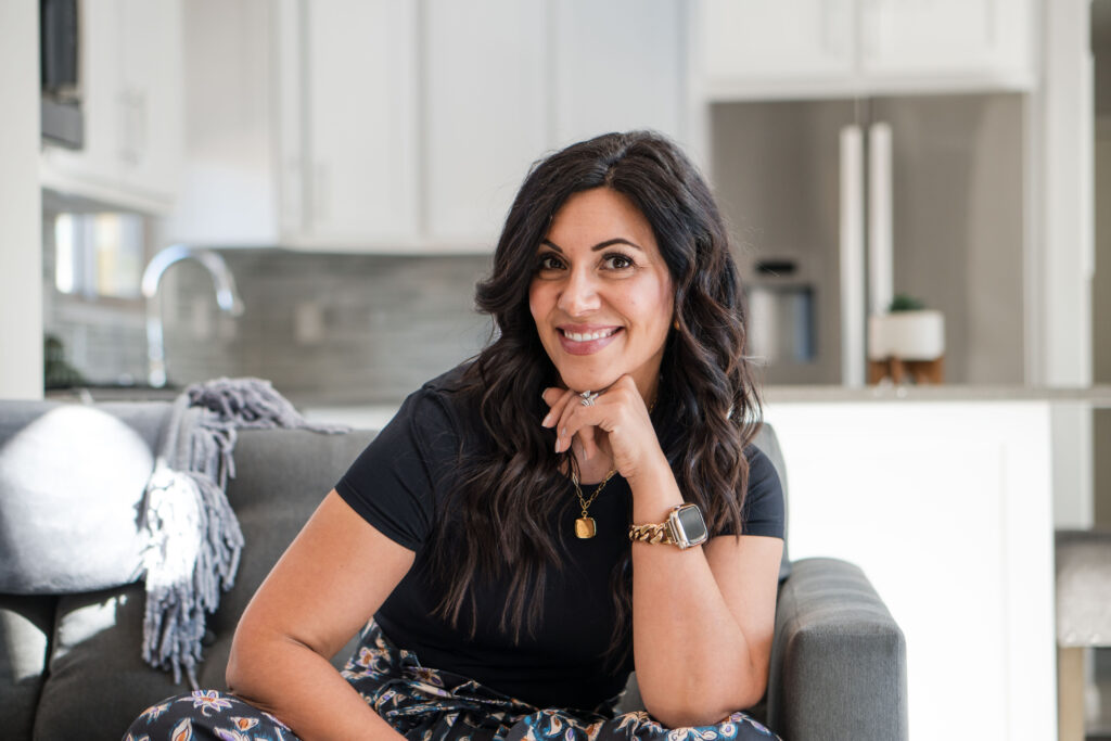 A dark-haired woman wearing a black t-shirt while sitting on the couch with the kitchen in the background. The title of the image is: Why-You-Need-to-Struggle-at-First-to-Succeed-Later-Stefanie-Gass