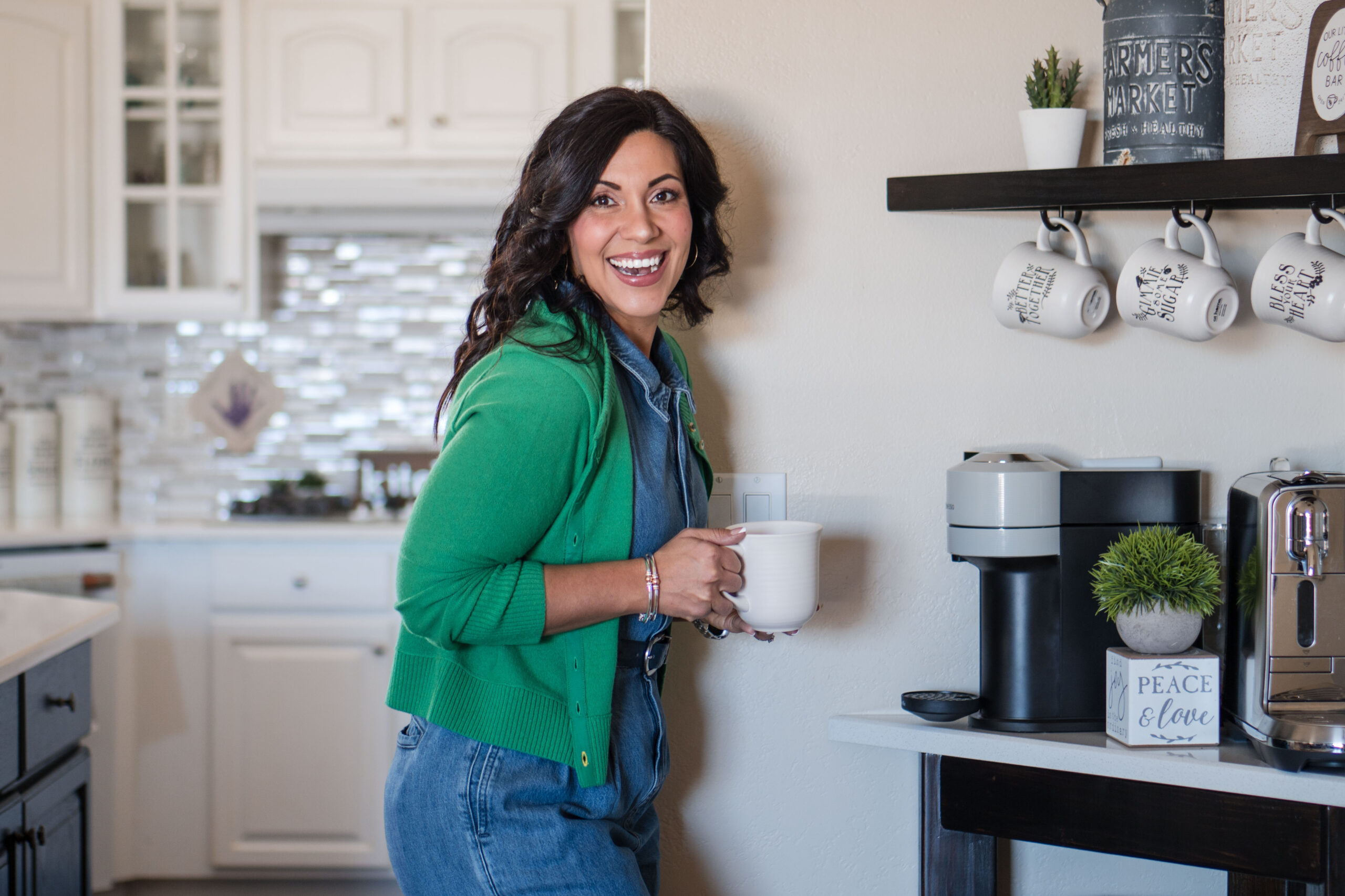 A dark-haired woman wearing a green sweater and blue jean pants is holding a white cup in her hand and smiling at the camera from the side. The image title is: CEO-Retreat-Recap-2024-Revenue-and-My-Strategic-Plan-for-2025-Stefanie-Gass