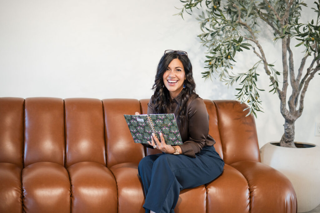 A dark-haired woman is sitting on a brown leather couch. She's wearing a brown silk blouse and blue jeans. She's reading from her black planner. The image title is: Redefining-Wealth-and-Well-Being-With-Patrice-Washington-Stefanie-Gass