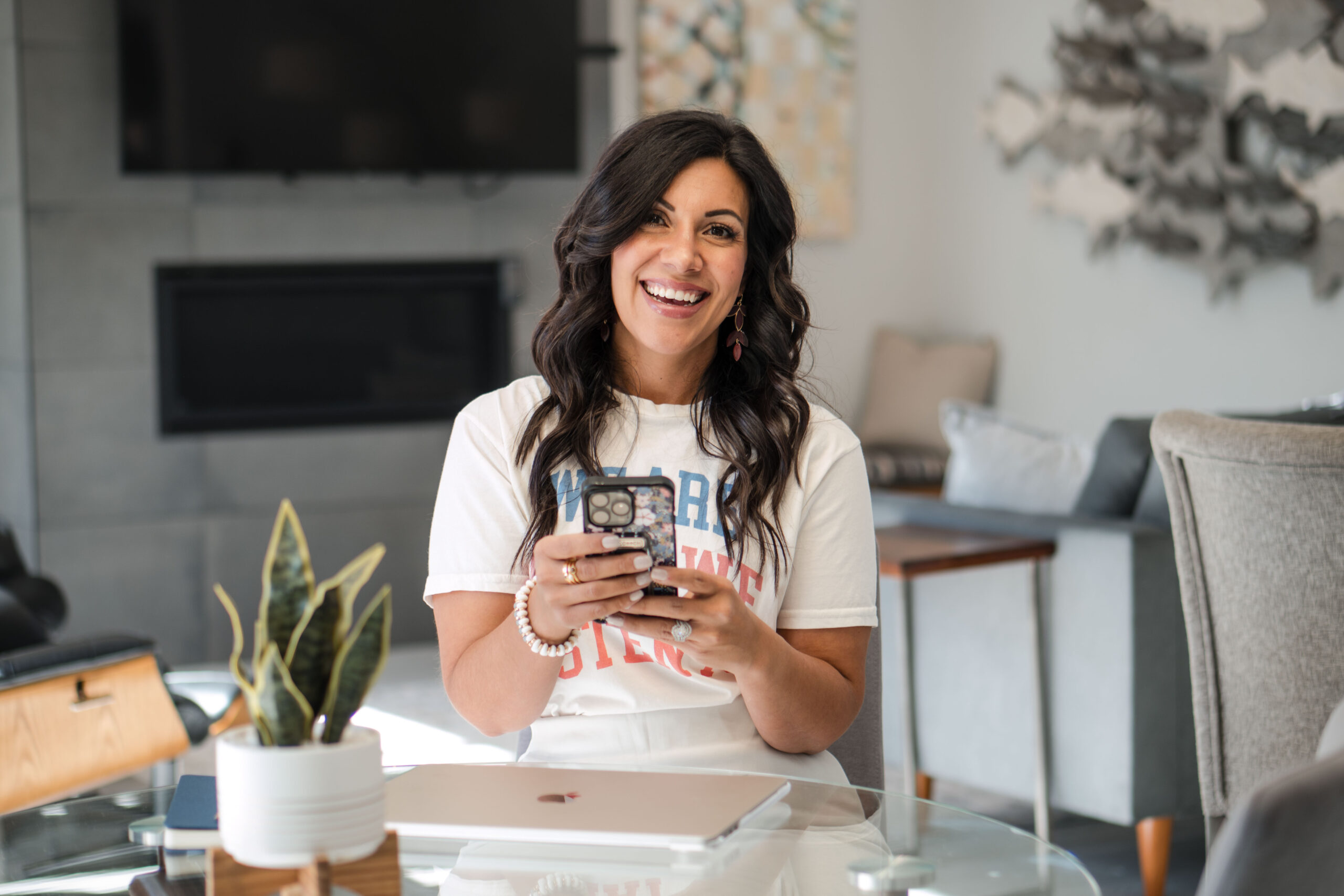 A dark-haired woman is holding her phone while smiling at the camera. She's wearing a graphic white t-shirt. Her white laptop is in front of her. The image title is: Three-Must-Dos-for-a-God-Led-Business-in-2025-That-Makes-Income-Stefanie-Gass